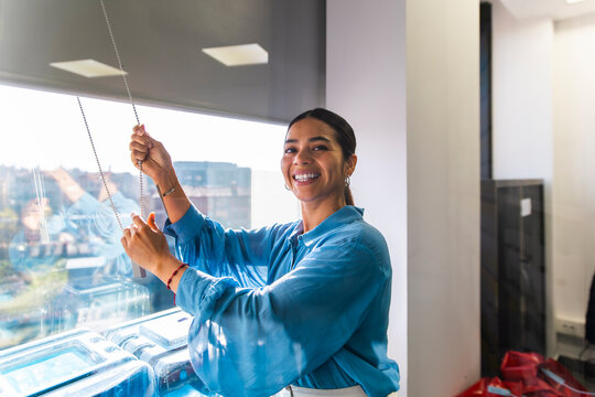 Smiling businesswoman opens the window blind in her sunny office