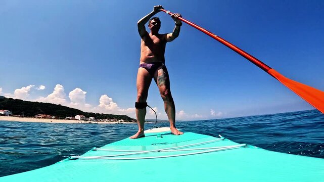 Slow-motion of a man who practices stand-up paddleboarding in the Mediterranean Sea along the coast of Sicily, the man is forty-two years old and of Caucasian origin