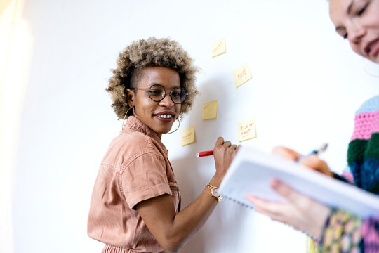 Businesswoman writing on adhesive notes while discussing with female colleague at home office