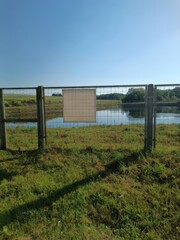 Fototapeta premium A body of water behind a chain-link fence under a bright blue sky, with a blank white sign, green grass in the foreground.