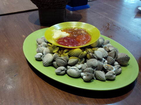 Fresh steamed cockle shellfish served with assorted dipping sauces on a green plate, representing authentic Southeast Asian seafood cuisine and restaurant dining.