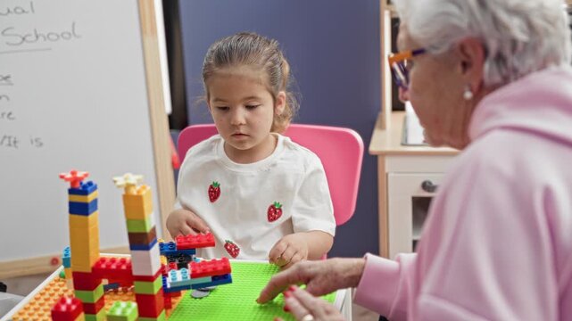 Young girl stacking toy blocks with hands while senior woman reaches to guide on a table in a school building; bonding calm.