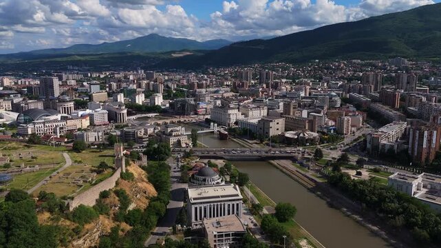 Aerial drone tracking shot following the Vardar River flowing through the urban center of Skopje, North Macedonia. Scenic city landscape with bridges and buildings at daylight