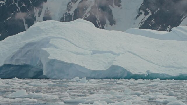 Large tabular iceberg sits amid fractured sea ice with steep snow covered peaks rising behind it in Antarctica