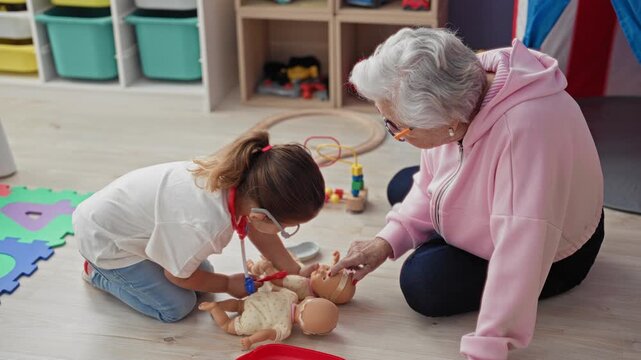 Girl uses toy stethoscope on baby doll while senior woman guides her hand in a playroom building; intergenerational tenderness.