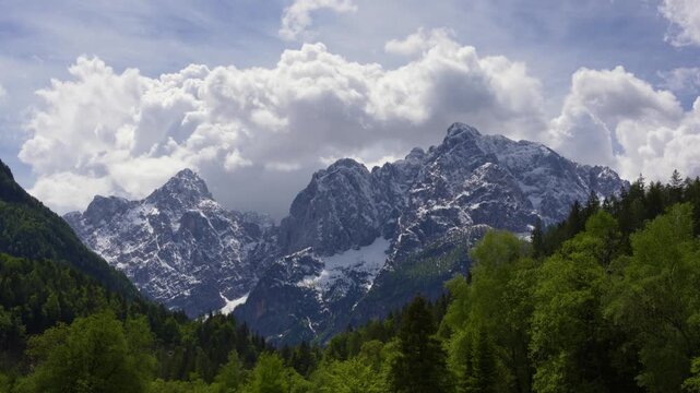 Snow-capped Julian Alps rise above green forest near Kranjska Gora, with dramatic clouds framing a bright alpine spring landscape.