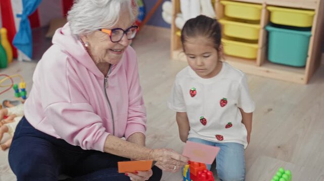 Woman in pink hoodie holds a flashcard to a little girl seated among toys in a daycare classroom; intergenerational learning calm.