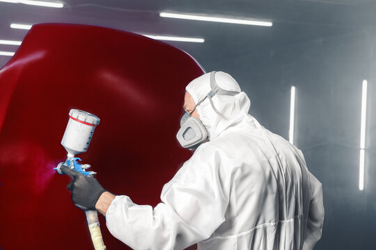 Automobile repairman painter in protective workwear and respirator paints part of car body in paint chamber using airbrush, close up. Restoration painting of car in the paint chamber service. 