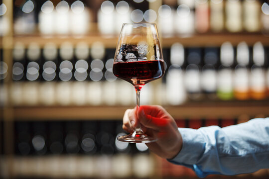 Close-Up of Sommeliers Hand Holding Glass of Red Wine Against Shelf of Assorted Bottles