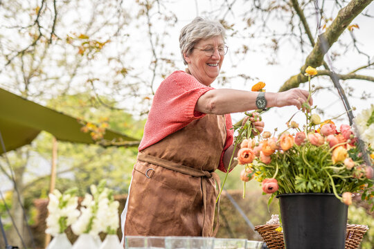 A cheerful senior woman in a brown apron smiling while arranging a colorful bouquet of ranunculus flowers at an outdoor table, celebrating creativity and joy in floristry.