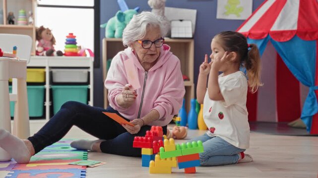 Senior woman holds flashcards and points while little girl covers ear and sits among blocks on a playroom floor; learning bonding warmth.