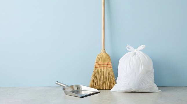 A classic broom metal dustpan and white trash bag standing against a soft blue wall background