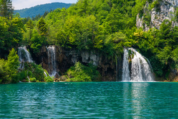 Fototapeta premium Vibrant turquoise lake mirrors lush green forest, with multiple cascading waterfalls flowing down rugged cliffs, creating breathtaking natural landscape. Plitvice Lakes is National Park in Croatia.