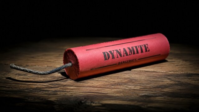 Close-up of a single red dynamite stick with printed label and gray fuse resting on a rustic wooden surface against a pitch black background with dramatic lighting