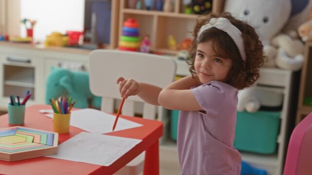 Girl hispanic child drawing with colored pencil at small red table in daycare building, leaning over paper surrounded by toys and crayons; calm concentration.