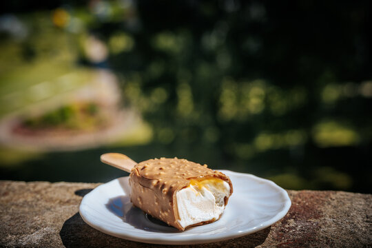 Close-up of a bitten caramel ice cream bar with creamy filling on a white plate outdoors in sunlight.
