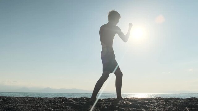 Slim lonely young man trains boxing and hand to hand combat punches on the seashore at sunset. Jab and uppercut in slow motion, outdoor workout, shadow boxing or shadow fighting, combat sports.