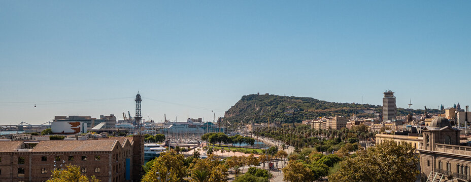 A panoramic view of Barcelona featuring the coastline, hills, and urban architecture under a clear blue sky. The scene captures the vibrant city life and natural beauty.