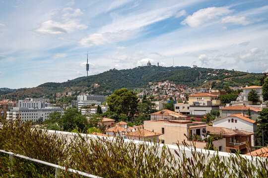 A panoramic view of Barcelona showcasing residential buildings and a green hillside. The iconic Collserola Tower is visible in the background under a clear blue sky.