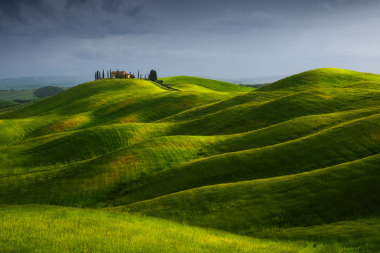 The Podere Staffolino sits on a rounded hilltop surrounded by deeply saturated green rolling wheat fields, a distant rain shower visible under a stormy sky. Crete Senesi, Tuscany, Italy.