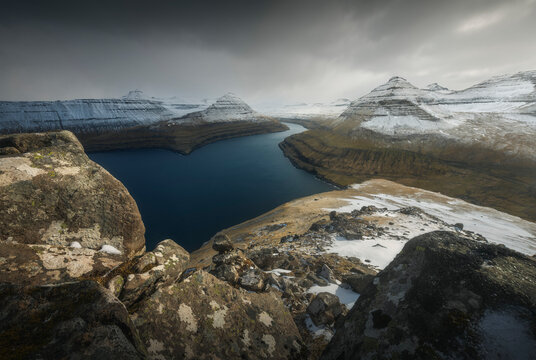 Snow-capped layered basalt mountains frame a deep narrow fjord viewed from above, Eysturoy, Faroe Islands.