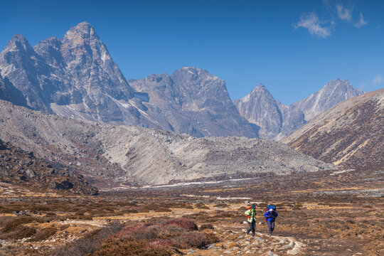 Two trekkers cross a wide, barren high-altitude plateau, the dramatic jagged grey ridgeline of Cholatse towering massively overhead in the midday light.