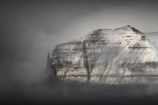 Snow-covered dramatic sea cliff emerges from dense fog over dark water, Faroe Islands.