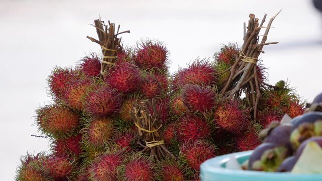 Piles of red rambutan in bunches with short stems sit on a market stall, with other fruit in a bowl in front. Exotic fruit.