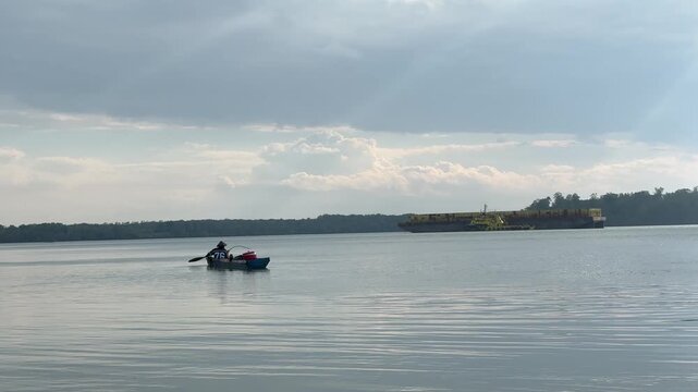 The silhouette of a fisherman angling while rowing across vast, calm waters beneath a cloudy sky. In the distance, yellow barges are moored in a peaceful bay, with the horizon framed by mangrove trees