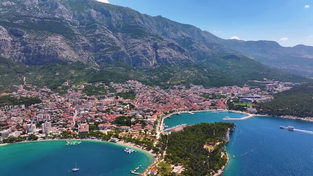 View of Makarska Croatia from above showing the coastline, buildings, and mountains. The scene includes water, trees, and roads. Bright sky with few clouds visible.