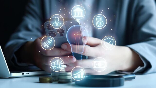 A person uses a smartphone, displaying technology and finance symbols in the air. Coins and a laptop are on the table. The focus is on the interaction with digital symbols and devices.