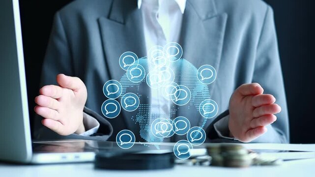 A business person sits at a desk with a laptop and stacks of coins nearby. They are using hand motions to interact with digital chat icons floating above the desk, showing conversation flow.