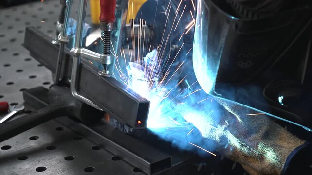 A engineer TIG welds metal structural parts, which are fixed with clamps. He wears a protective mask and gloves to avoid injury from hot sparks