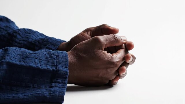 Close-up of clasped hands of a Black person resting on a white surface, wearing a blue textured long-sleeve shirt, interlocked fingers in a gesture of prayer or meditation.