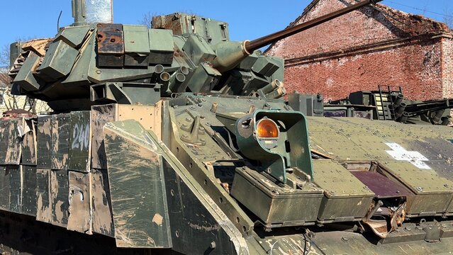 Damaged infantry fighting vehicle with explosive reactive armor blocks, a white cross marking, and a smashed headlight parked near a brick wall.