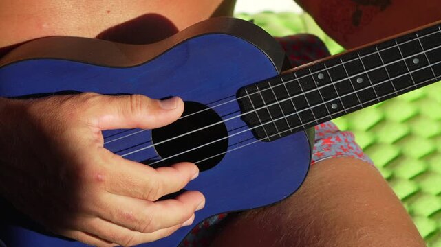 Ukulele, playing, man strumming blue instrument outdoors in sunlight.