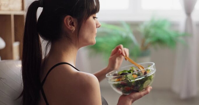Woman eating healthy salad at home. Seated in sportswear near a bright window, she holds a glass bowl, focusing on nutrition and wellness after a workout. Clear concept of a healthy lifestyle.
