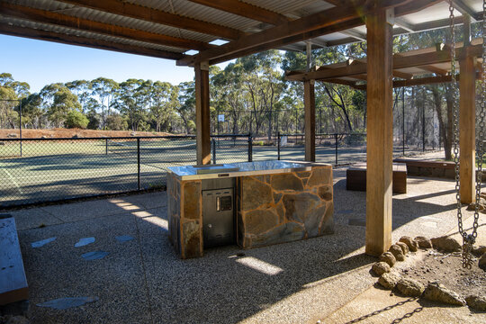 A public barbecue facility featuring a hotplate and a  stone-clad base sits sheltered beneath a pavilion. Outdoor recreational space overlooks a fenced tennis court amenities. Eynesbury, VIC Australia