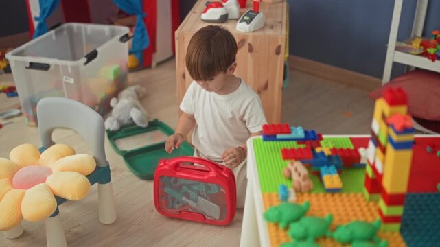 Boy sitting on playroom floor holds red toy tool case with hand among scattered blocks and storage bin; quiet concentration.