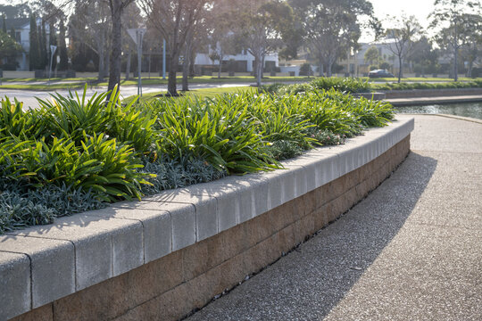 A long, curving raised garden bed built from textured interlocking retaining wall blocks spans alongside a public concrete footpath. A landscaped buffer between walking path and a residential street