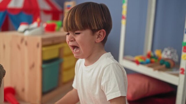 Boy crying with open mouth in a colorful playroom near a tent and toy shelf, reaching toward scattered toys and a pillow; tantrum sadness.