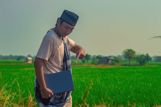 Asian Muslim man wearing a peci hat and sarong while holding a laptop and pointing down at a green rice field, presenting smart agritech business.