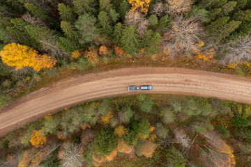 Naklejka premium Overhead drone view of an SUV with a camping trailer driving on a curving dirt road surrounded by dense autumn woods.