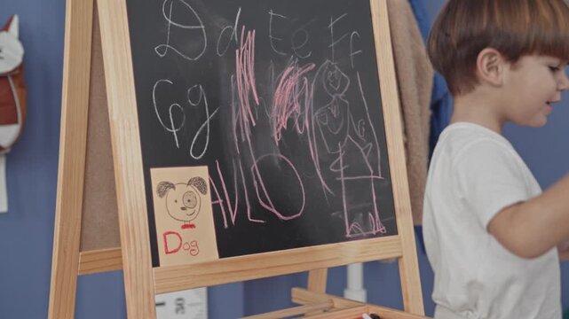 Boy in white t shirt drawing letters and pink scribbles on a freestanding chalkboard easel, holding pink chalk and touching board in a studio; curiosity discovery play.