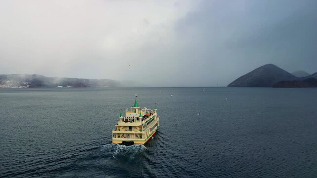 Drone shot of a classic passenger ferry cruising across Lake Toya towards snow-dusted islands under a cloudy winter sky in Hokkaido, Japan.