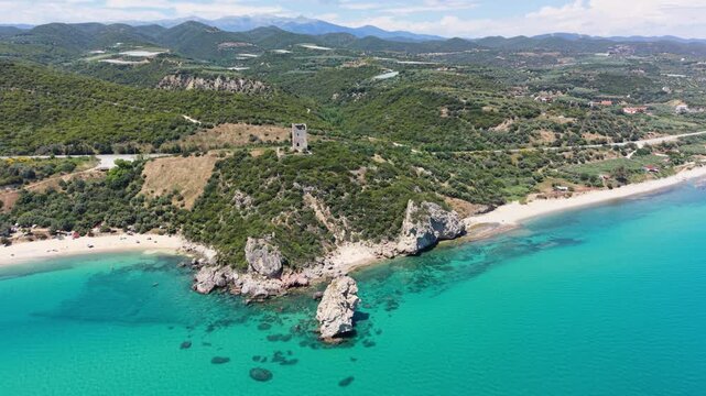 Drone orbits around Apollonia Tower and Paralia Folias in Greece, revealing turquoise Ionian waters, a rocky shoreline, sandy beach, and the surrounding coastal hills.