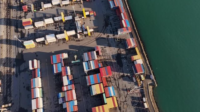 Colorful shipping containers stacked at industrial cargo port, rail tracks, small crane, teal harbor water, dock yard, shadows, Wellington, North Island, New Zealand. Aerial drone top down shot