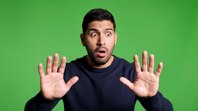 Shocked young man with a dark beard wearing a navy blue sweater holding both hands up with palms facing outwards in a defensive gesture looking wide-eyed directly at the camera against a solid green b