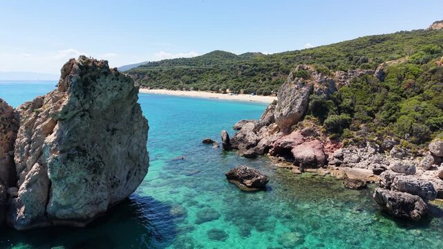 Drone passes between striking sea rocks and continues toward the beach at Paralia Folias on the Ionian coast of Greece, revealing turquoise water and a rugged shoreline.
