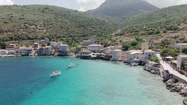 Aerial view of coastal village Limeni on Mani peninsula, waterfront old stone house towers surrounded by lush green mountains, moored fishing boats in turquoise waters on a sunny day